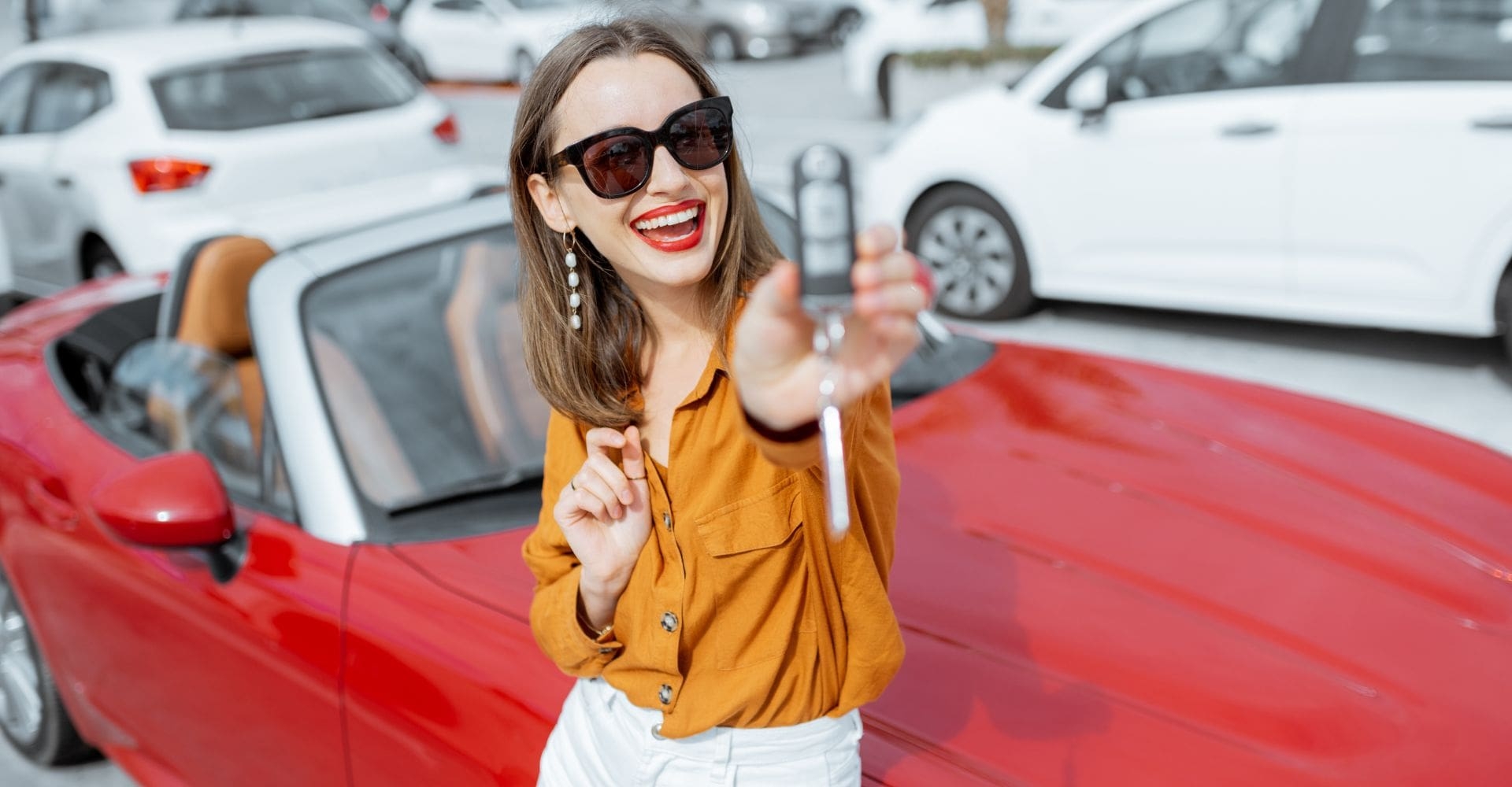 woman wearing sunglasses holding up the keys to her red rental car in excitement 