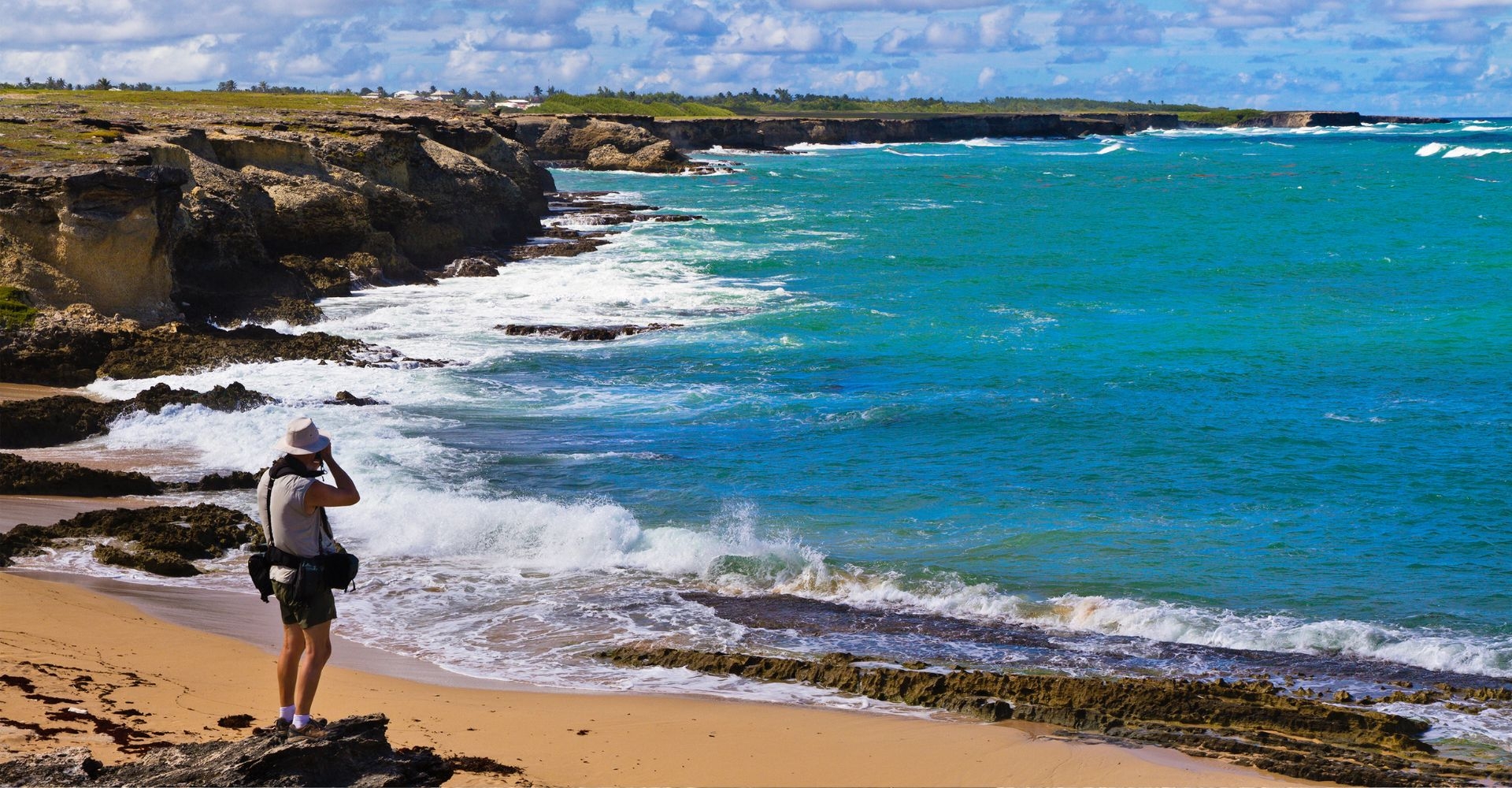 a man wearing a sun hat and protective layers taking pictures on the coast on a windy day in Barbados