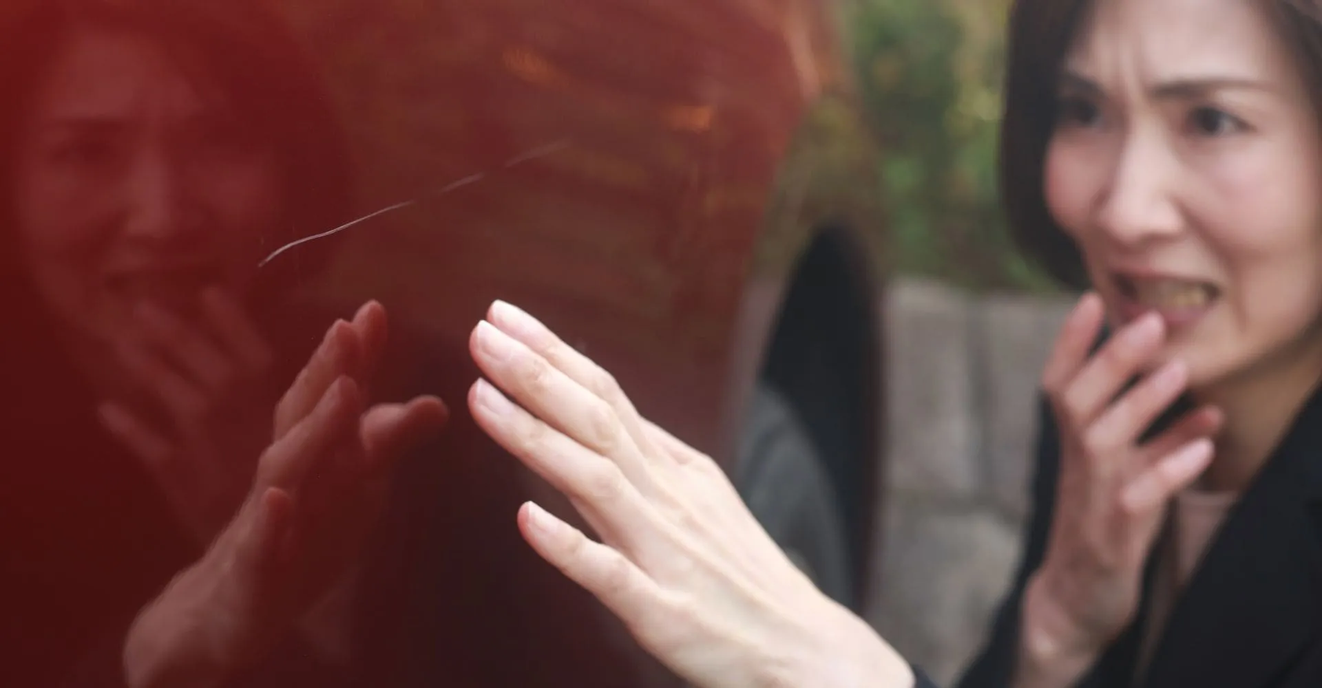 A woman stands beside a red car, her hand resting on the door as she examines a scratch on the vehicle