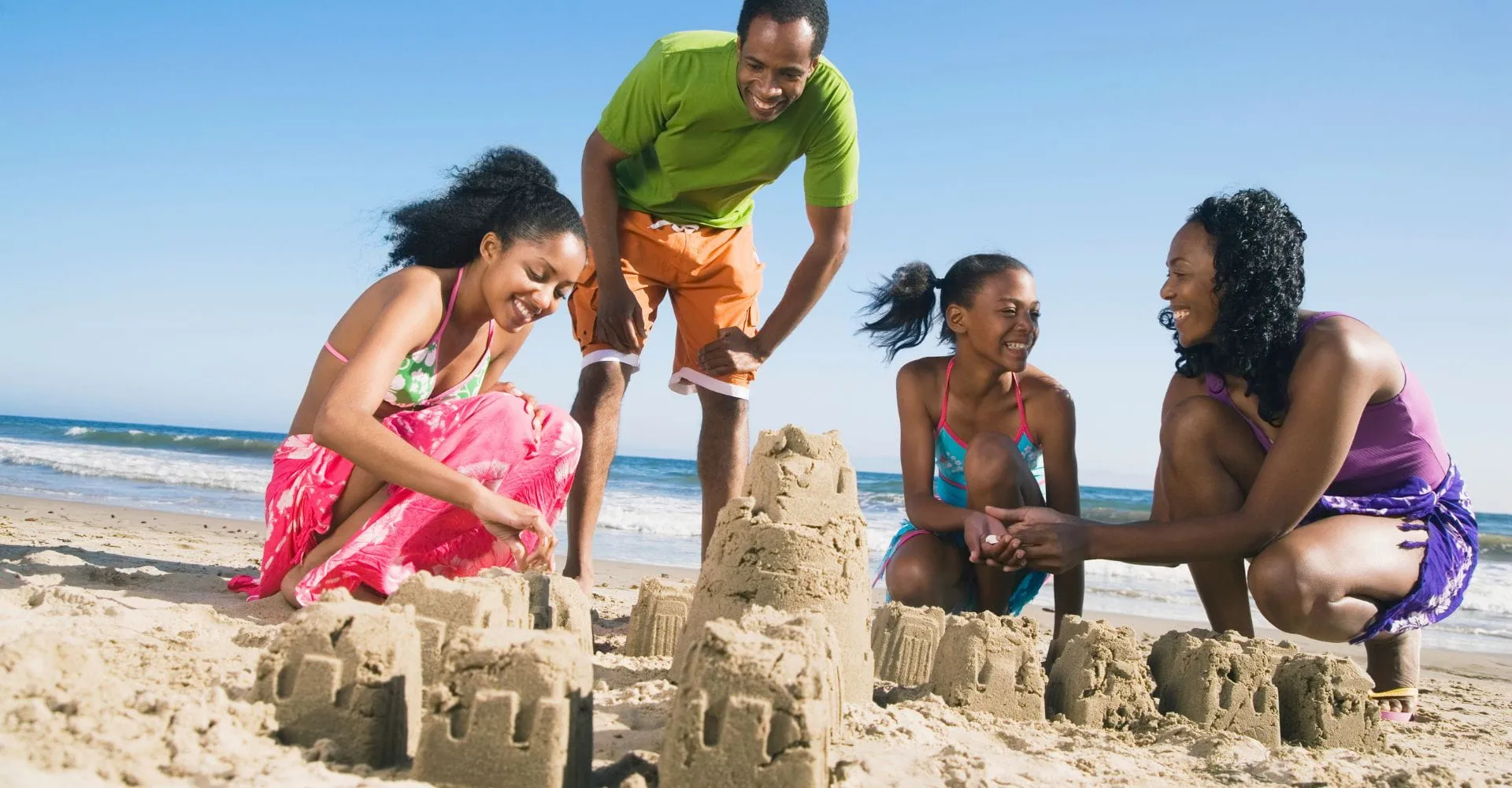 A joyful family of four builds sandcastles on a sunny beach. The clear blue sky and gentle waves set a cheerful, relaxed tone.