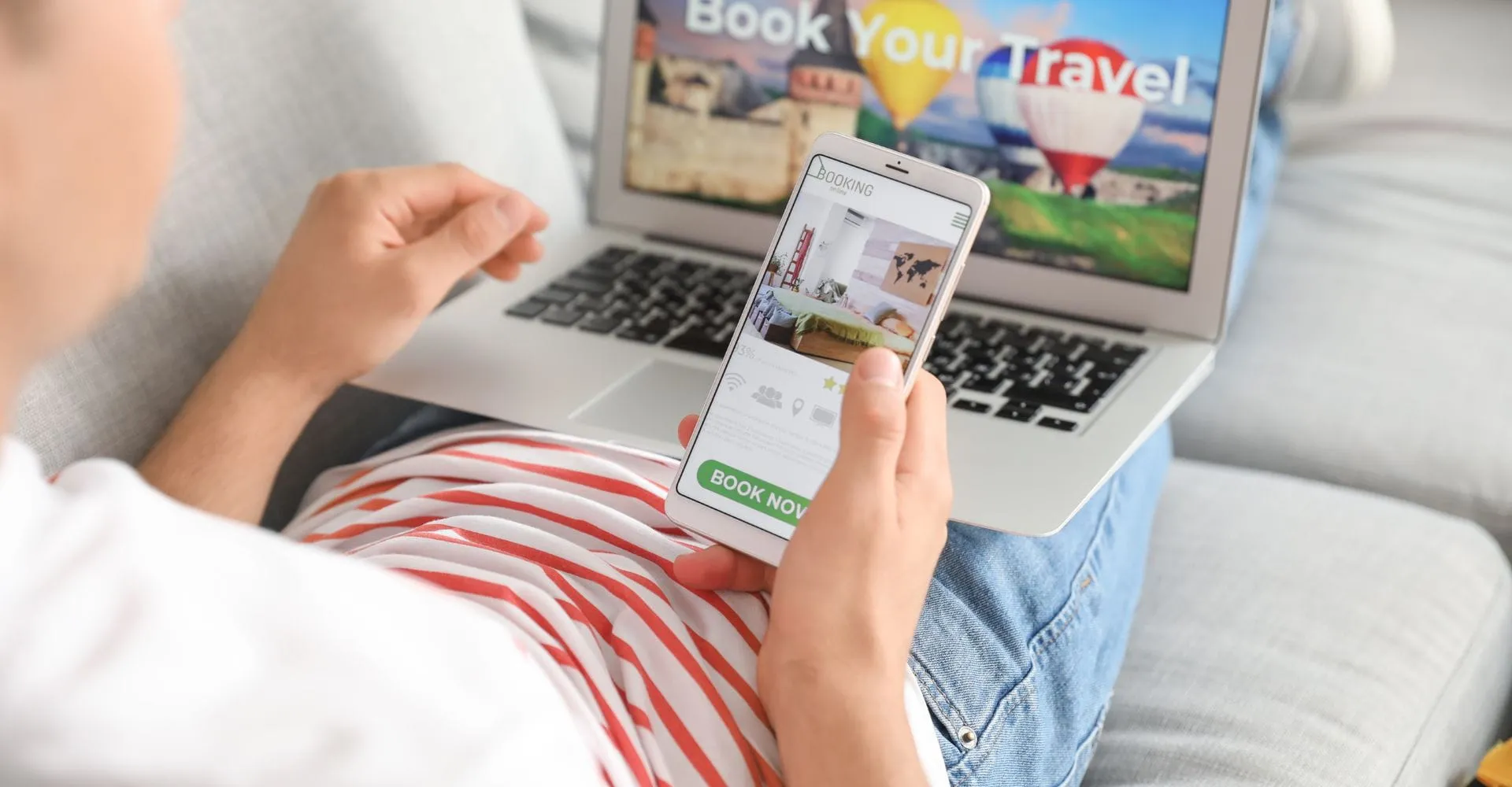 Person holding a smartphone and browsing accommodation options, with a laptop displaying "Book Your Travel" in the background.