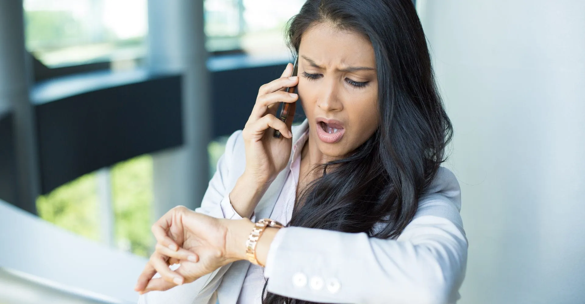 A woman in a white blazer looks stressed while checking her watch and talking on the phone. She is standing indoors, with large windows in the background.