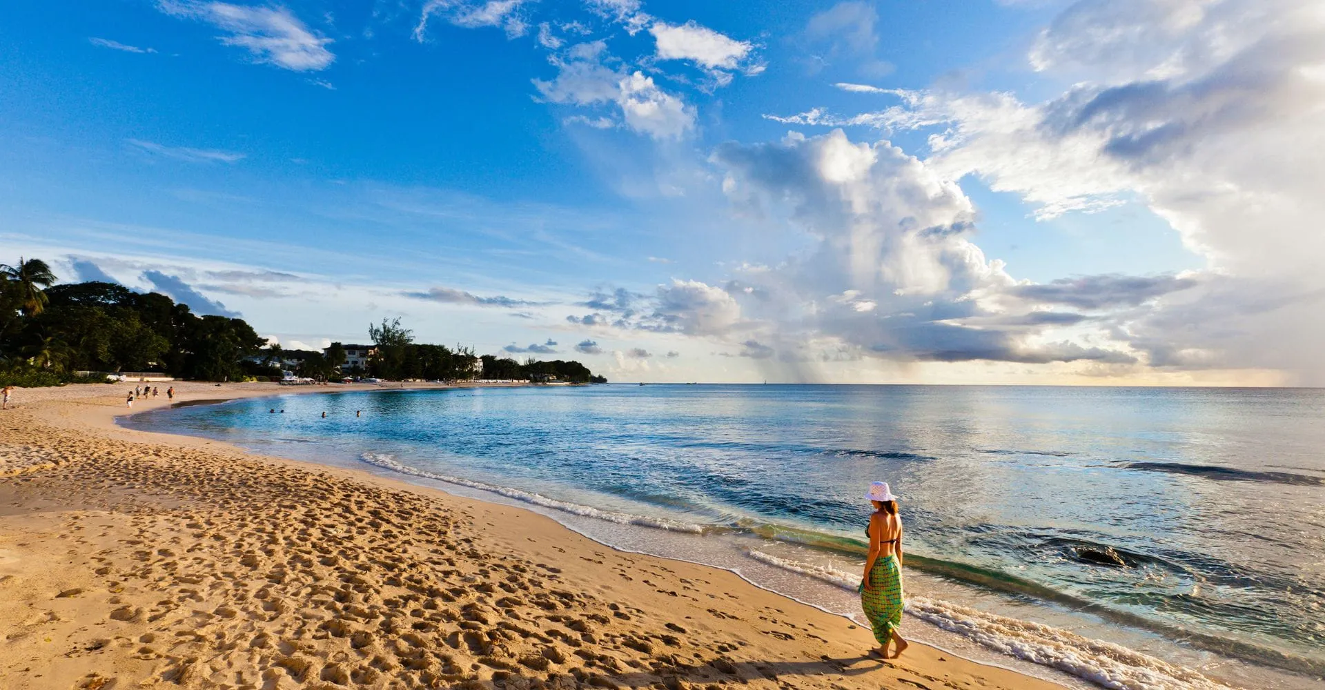 A person in a hat walks along a tranquil, sandy beach at sunset, with gentle waves and a vibrant sky with clouds, creating a serene atmosphere.