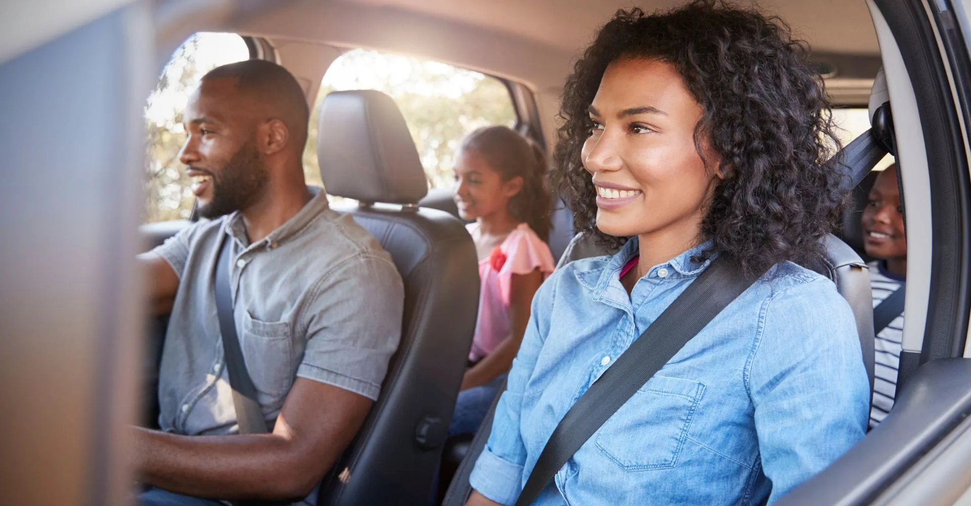 A smiling family of four sits inside a car. Parents are in the front seats with the father driving, and two children are in the back. Sunlight filters through the windows.