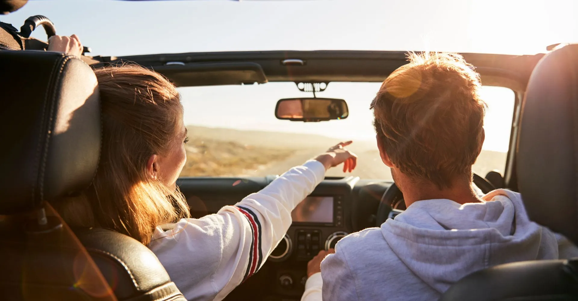 A woman and man drive in a convertible on a sunny day. She points ahead, smiling, as they enjoy the open road. The atmosphere is carefree and joyous.