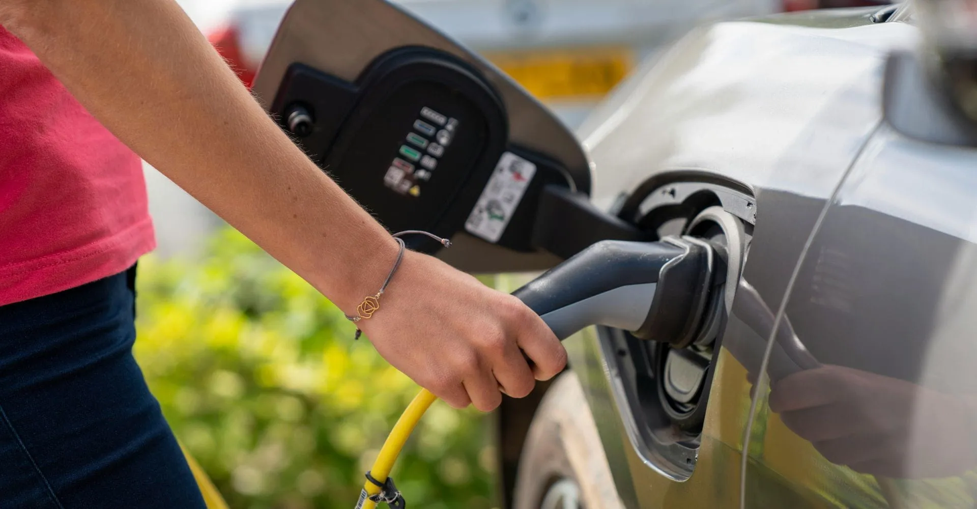 A person plugs a charging cable into a gray electric car, reflecting a focus on eco-friendly transport. Bright daylight suggests a positive tone.