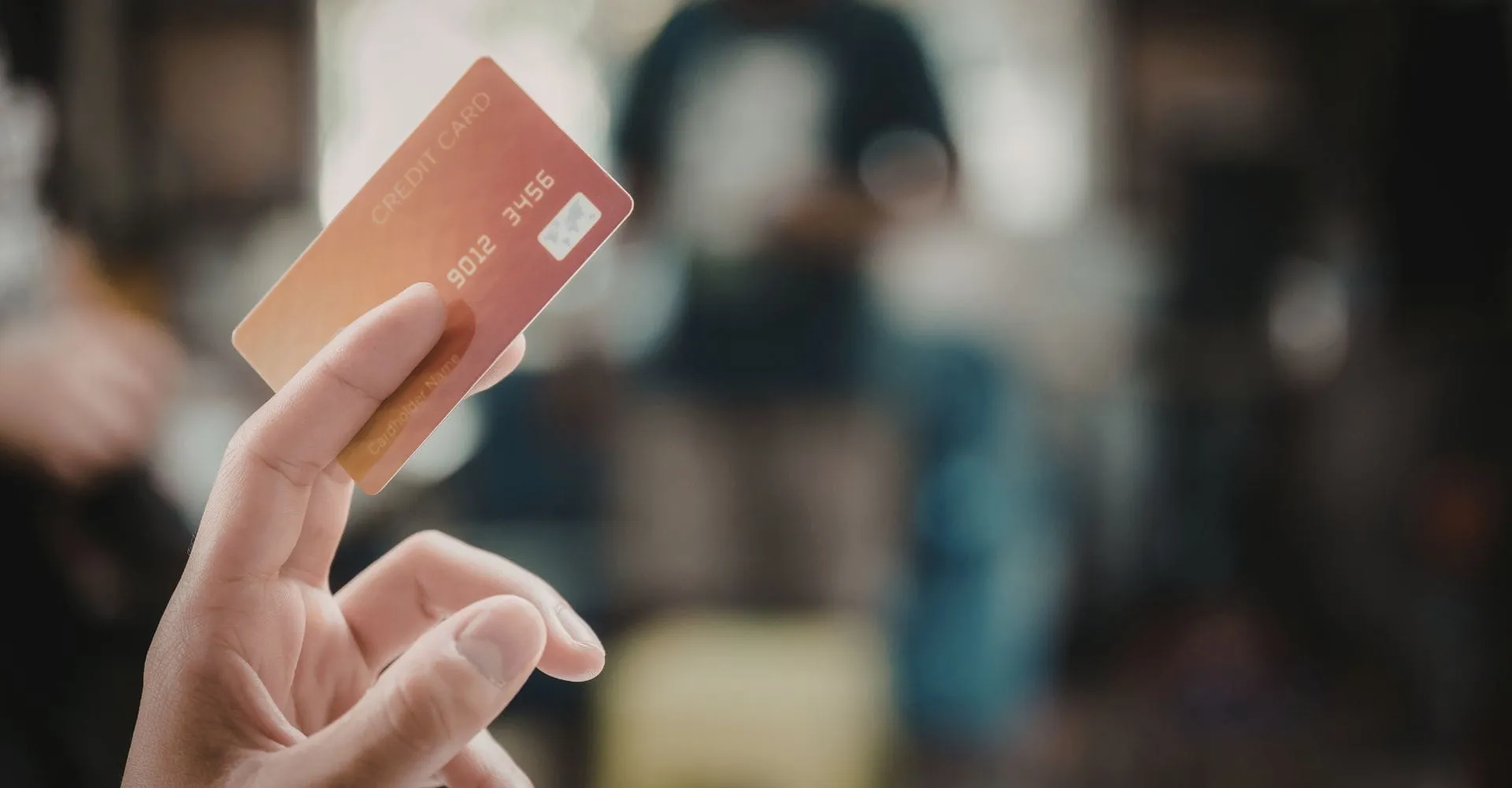 A hand holds an orange credit card between thumb and index finger, with a blurred background of people, conveying a sense of modern commerce or online shopping.