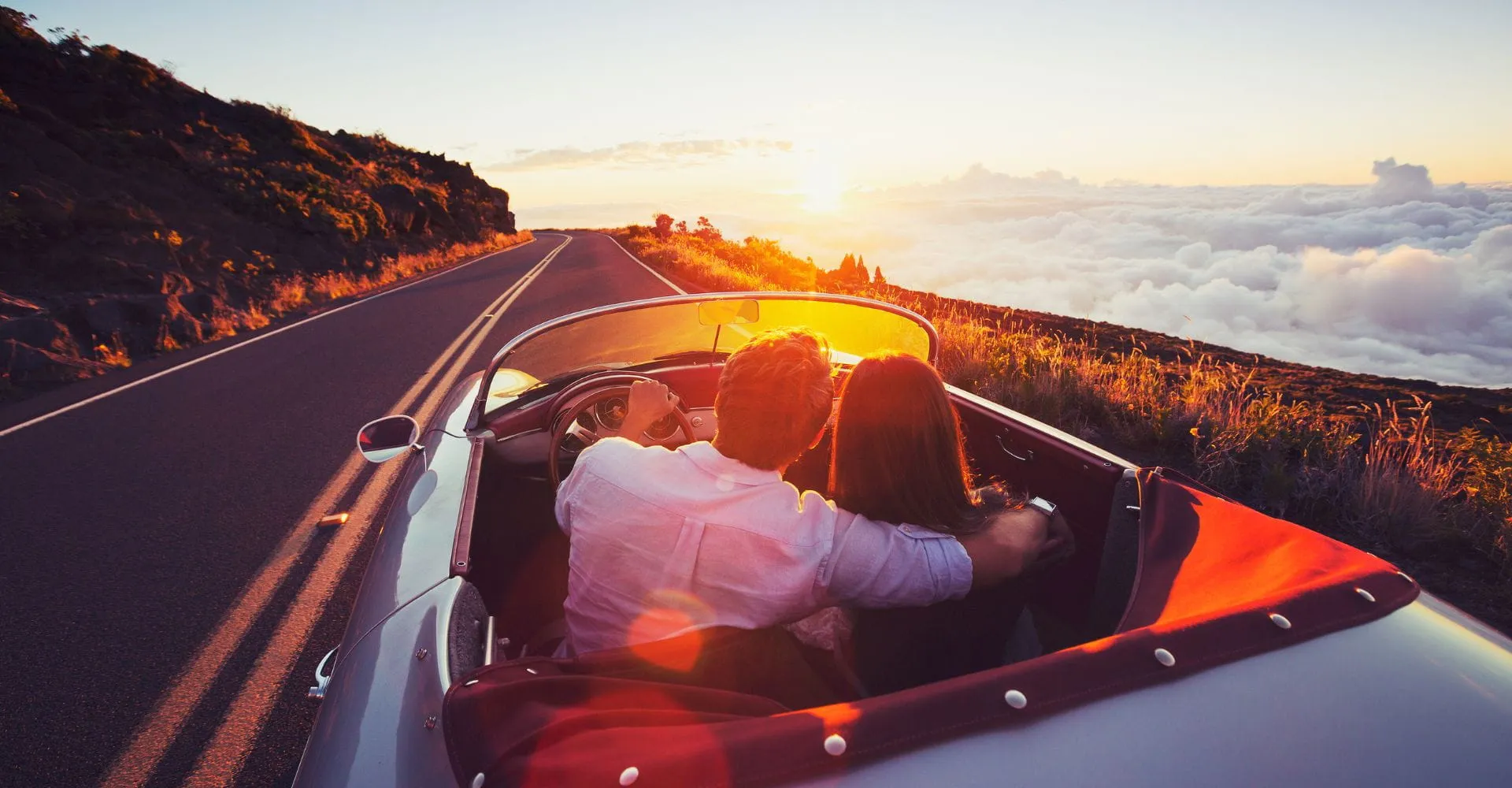 A couple sits in a convertible driving along a scenic road at sunset, with the sky painted in warm hues and a blanket of clouds below, evoking romance.