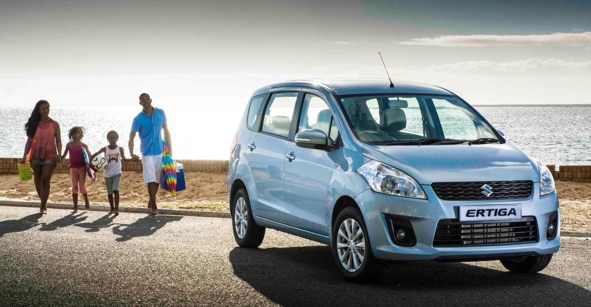 A family of four walks along a sunny beach road, holding colorful items. A blue Suzuki Ertiga is parked nearby, evoking a sense of leisure and joy.