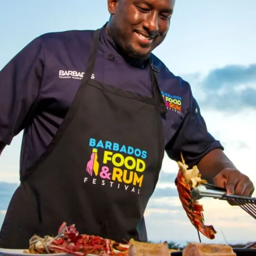 A man wearing an apron grills food on a barbecue, surrounded by outdoor greenery