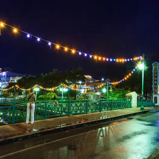 A brightly lit bridge spans a river at night, reflecting lights on the water's surface
