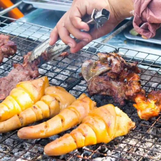 A person grilling meat outdoors, focused on cooking over a charcoal grill with smoke rising
