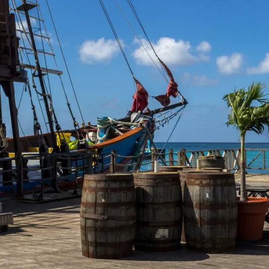 A wooden deck featuring barrels and a sailboat moored nearby, set against a serene waterfront backdrop