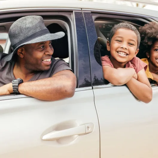 A family occupies the driver's seat of a car, with two adults in front and two children in the back, sharing a joyful moment.
