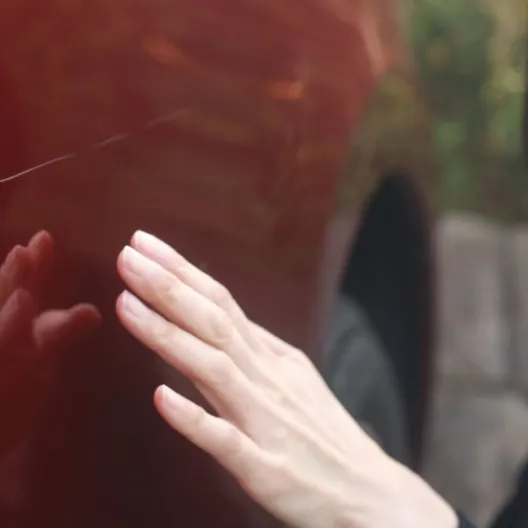 A woman stands beside a red car, her hand resting on the door as she examines a scratch on the vehicle