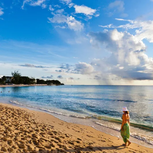 A person in a hat walks along a tranquil, sandy beach at sunset, with gentle waves and a vibrant sky with clouds, creating a serene atmosphere.