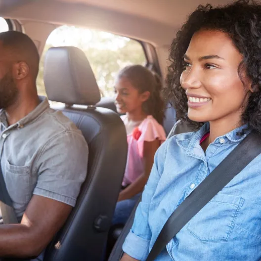 A smiling family of four sits inside a car. Parents are in the front seats with the father driving, and two children are in the back. Sunlight filters through the windows.