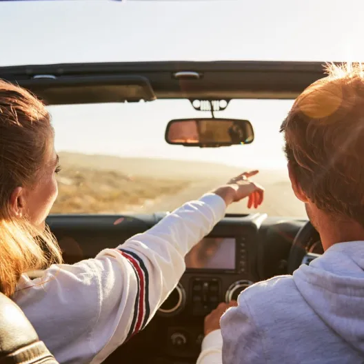 A woman and man drive in a convertible on a sunny day. She points ahead, smiling, as they enjoy the open road. The atmosphere is carefree and joyous.