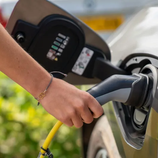 A person plugs a charging cable into a gray electric car, reflecting a focus on eco-friendly transport. Bright daylight suggests a positive tone.