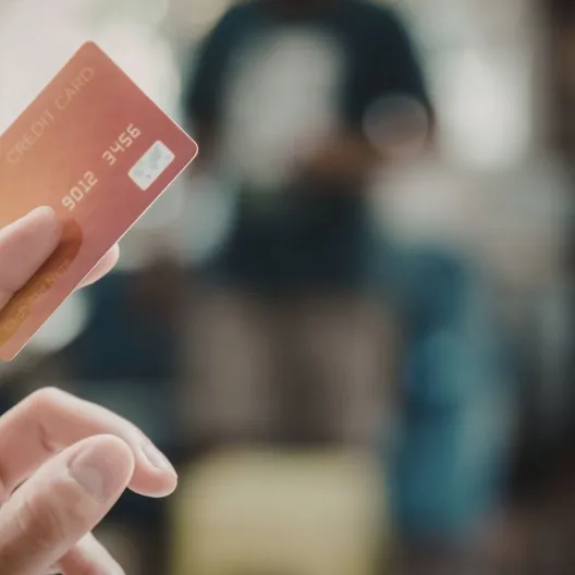 A hand holds an orange credit card between thumb and index finger, with a blurred background of people, conveying a sense of modern commerce or online shopping.