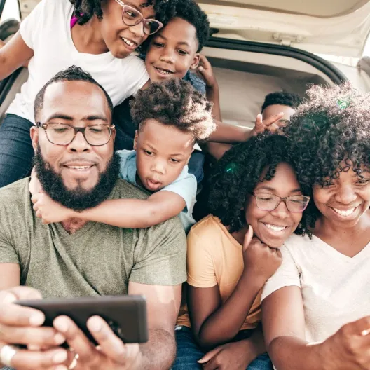 A joyful family of seven sits in the back of an open car, smiling and looking at smartphones. The scene conveys warmth, happiness, and togetherness.