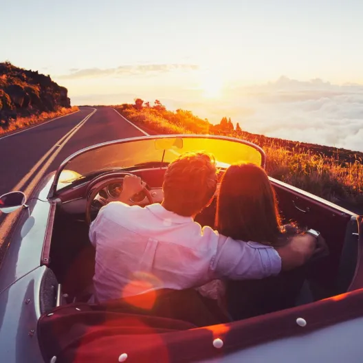 A couple sits in a convertible driving along a scenic road at sunset, with the sky painted in warm hues and a blanket of clouds below, evoking romance.