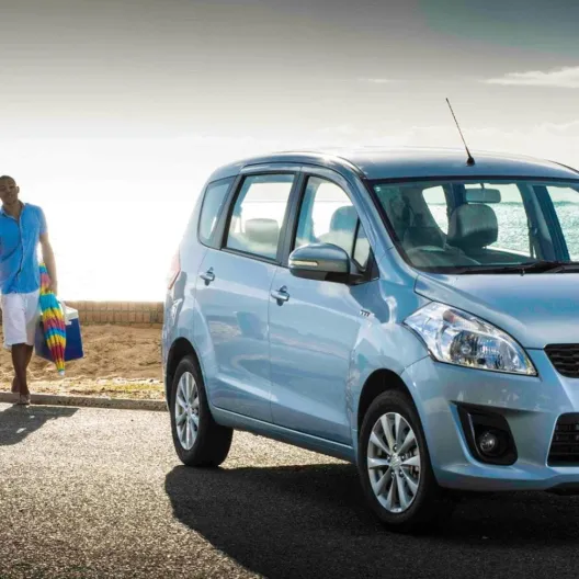 A family of four walks along a sunny beach road, holding colorful items. A blue Suzuki Ertiga is parked nearby, evoking a sense of leisure and joy.